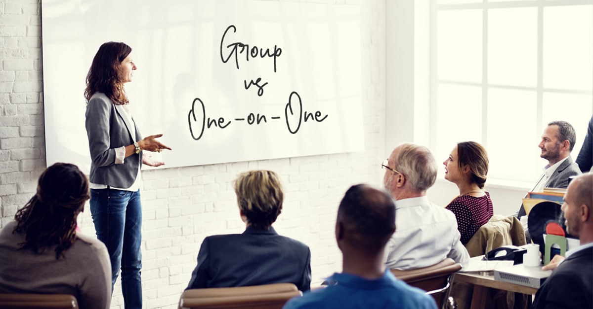 Female teacher at a whiteboard with the text "Group vs One-on-One" addressing a classroom of adults.