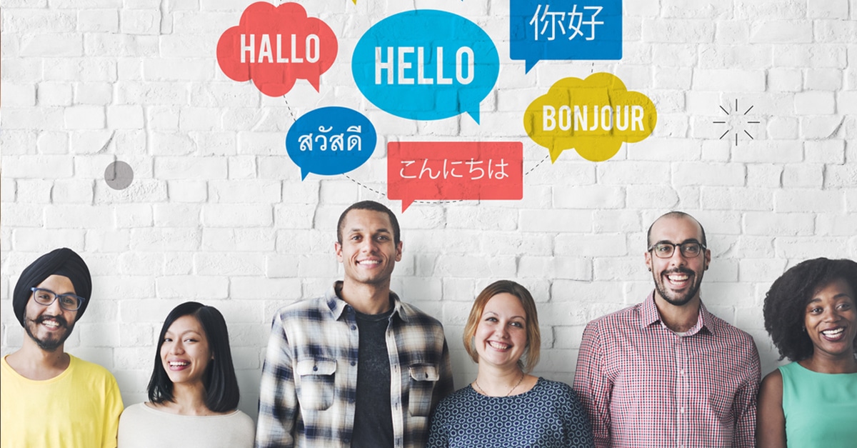 Six adults standing in a row looking at the camera with their backs to a white brick wall with multicolored text bubbles above them showing the word "hello" in different languages