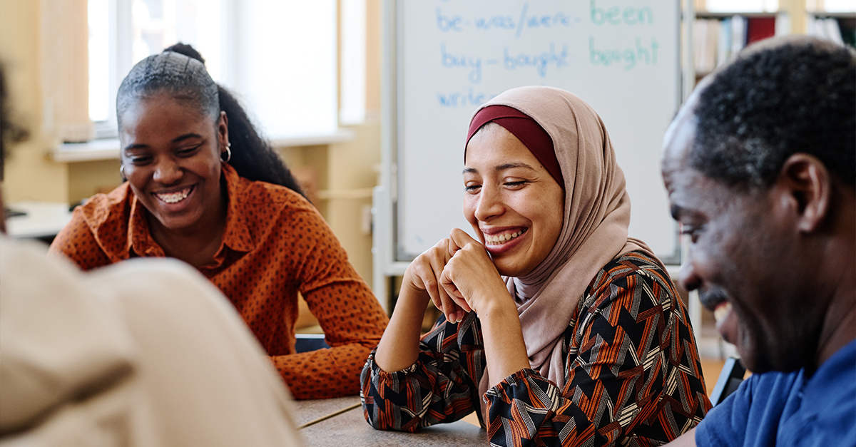 Group of modern immigrants sitting at table having fun laughing at something funny during english lesson