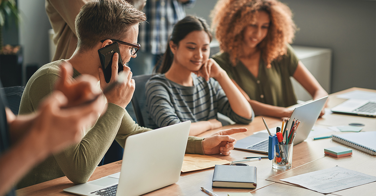 Multi-language workforce, man on phone at conference table next to two women looking at laptop