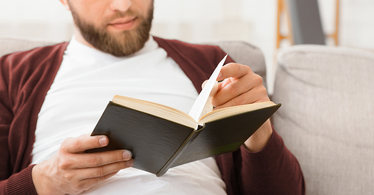 A man is seated on a couch, engrossed in reading a book. The man appears focused and relaxed, surrounded by a quiet atmosphere. | Best Books for Improving English Fluency Blog