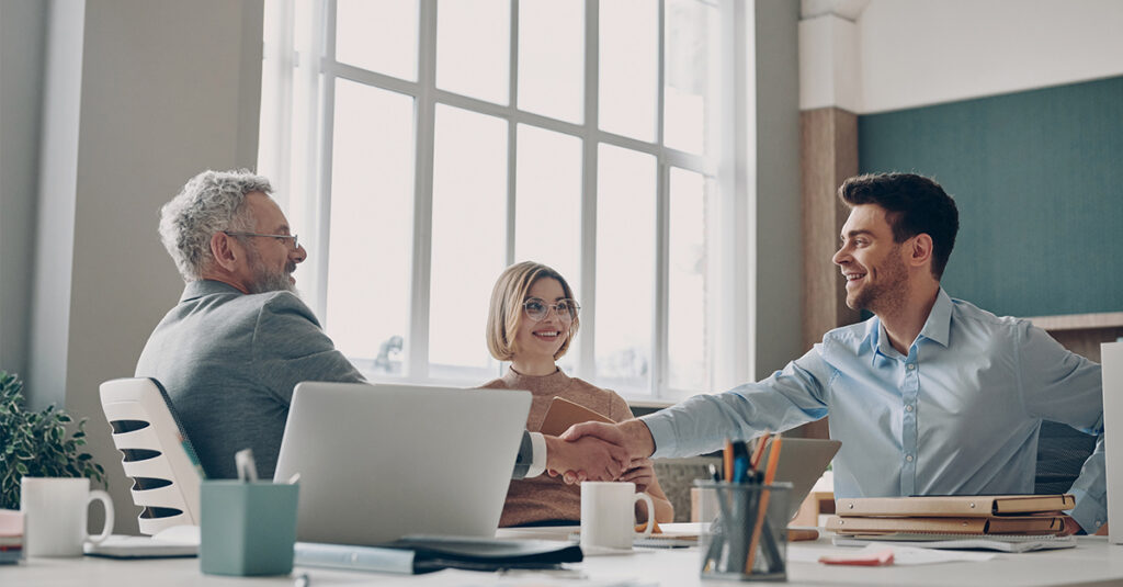 Business people shaking hands and talking while sitting together at an office desk, illustrating the blog topic "Why You Should Take a Spanish for Business Course — Even If You Already Speak the Language"