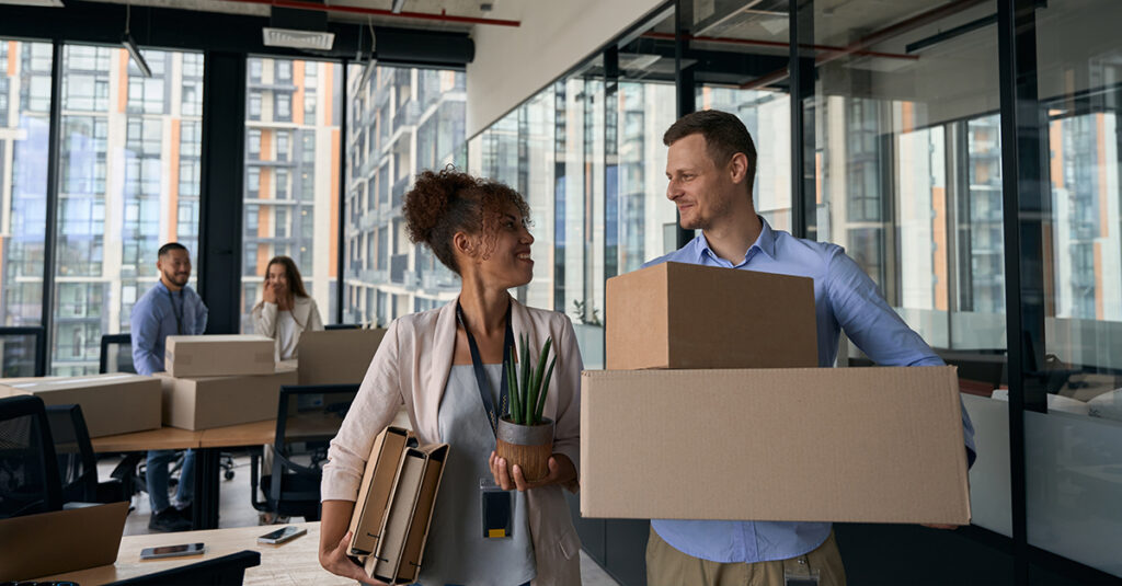 Professional woman and man carrying boxes, files, and a plant, preparing for relocation, Illustrating the blog topic "Corporate Relocation Packages: Why Language Training is the Missing Piece"