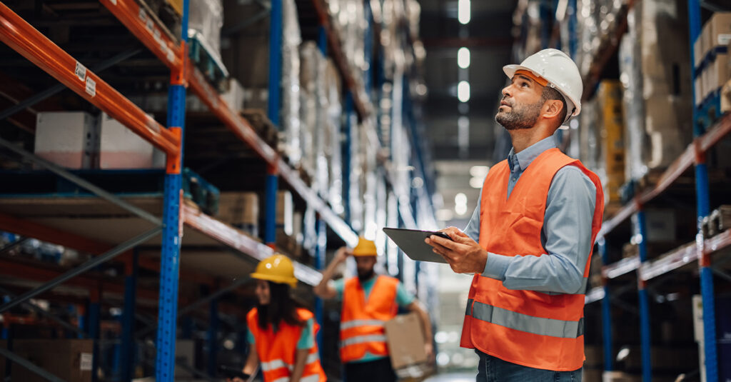 Warehouse manager holding digital tablet, looking up at shelves while checking inventory with his team in the background | Essential Spanish Vocabulary for Warehouse Work