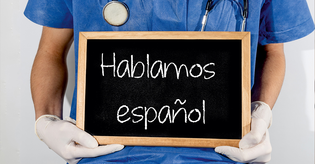 Closeup of a woman in scrubs with a stethoscope around her neck, sitting on a stool and holding a chalkboard with "Hablamos Español" to illustrate the blog topic "Why Clinics and Healthcare Practices Should Invest in Medical Spanish Training"