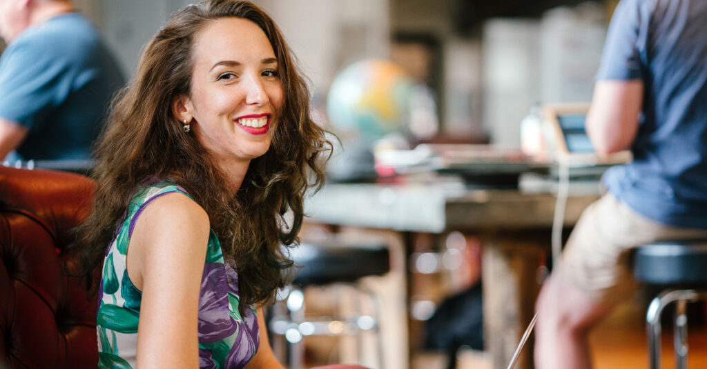 Portrait of a young and attractive European woman working on her laptop computer as she sits in a plush red armchair in a coffee shop or office during the day illustrating the blog topic of How to Integrate Expats into Your Company
