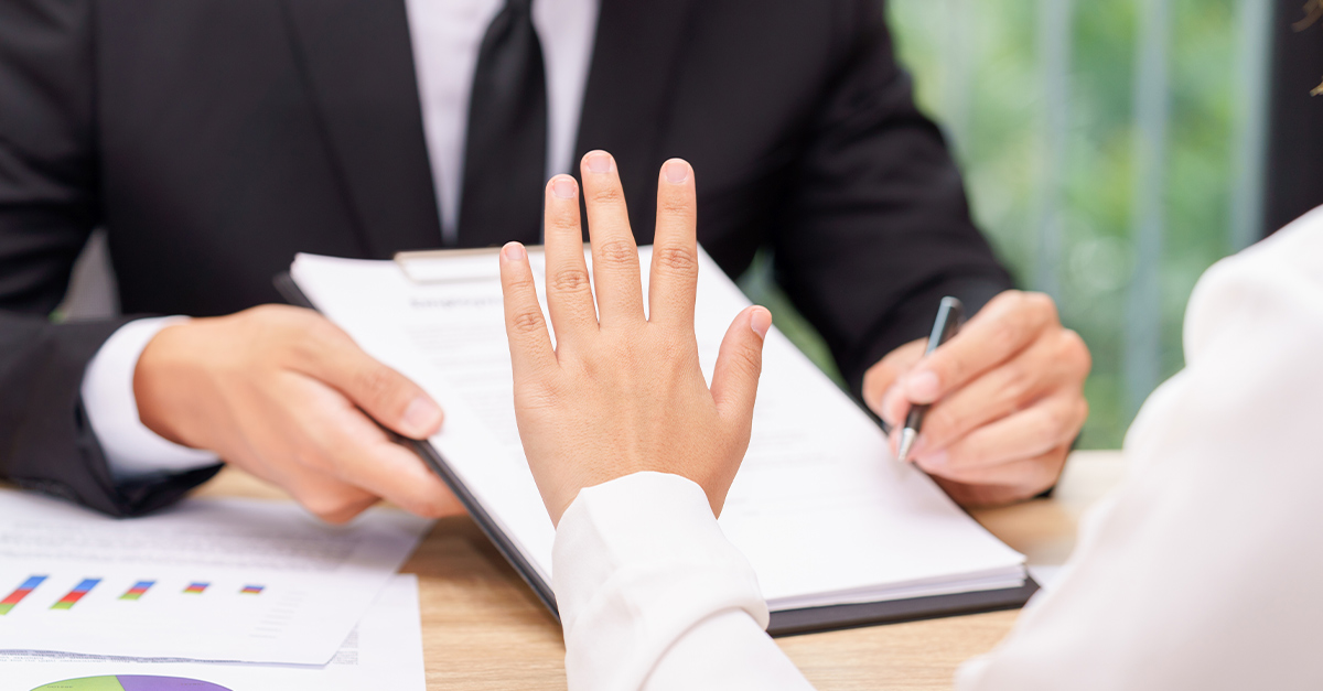 Woman's hand gesturing "no" or "hold on" to a man holding out a clipboard and pen across a desk