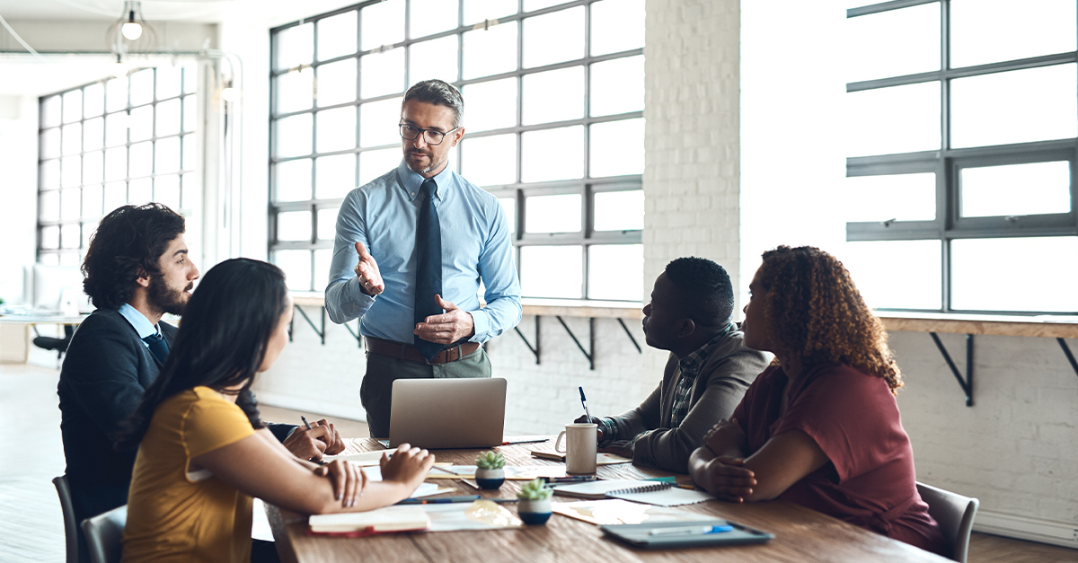 Bilingual manager leading a meeting, standing at a table of co-workers