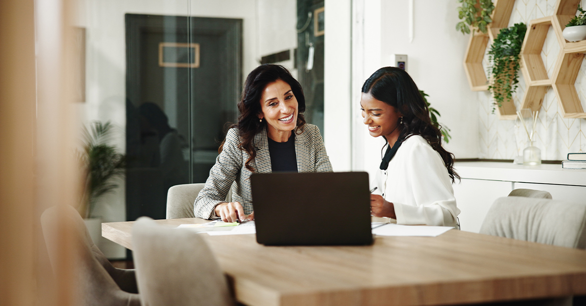 Smiling HR manager assessing female employee on her language skills at a wooden table in an office with an open laptop and paperwork.