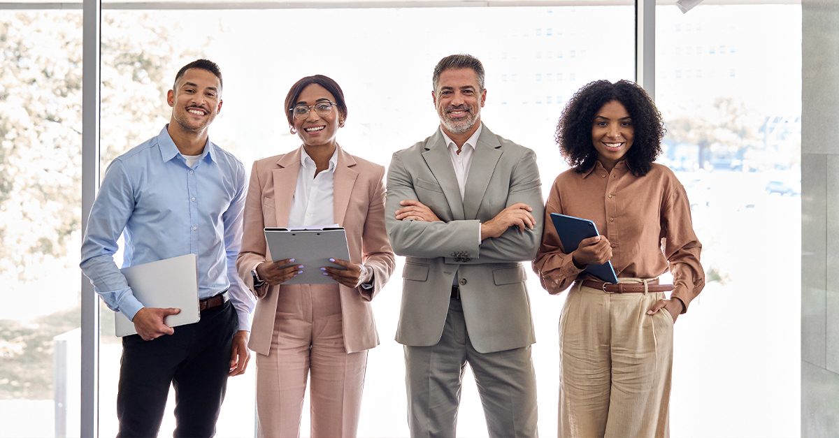 Four international professionals standing in front of windows in a bright office smiling at the camera, illustrating the blog topic Enhancing Cross-Cultural Communication in International Teams