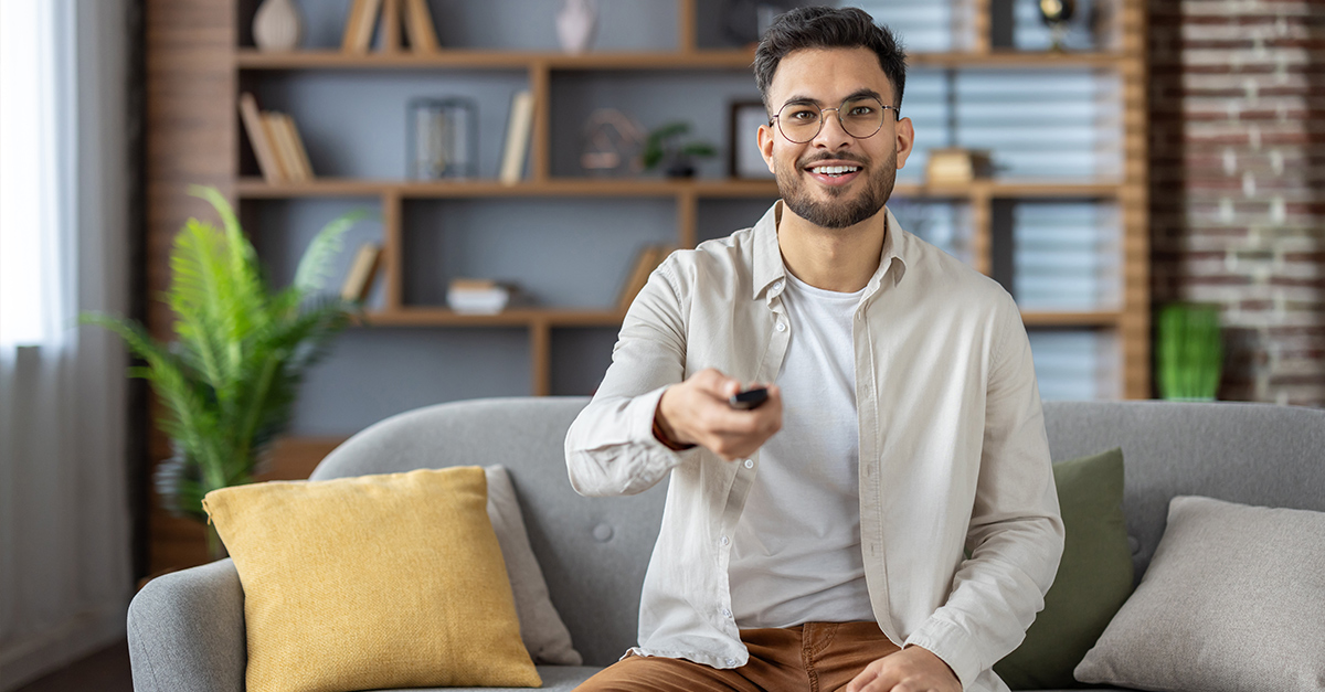 Man relaxing on sofa with remote control, enjoying leisure time in modern living room. Smiling and looking at camera, embodying comfort and relaxation while watching TV at home, illustrating the blog topic Mastering English Vocabulary through TV Shows