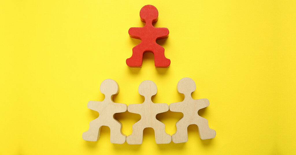 One red wooden block of a human figure above three plain wooden human figures on yellow background, illustrating the blog topic What's Your Language Learning Personality Type?