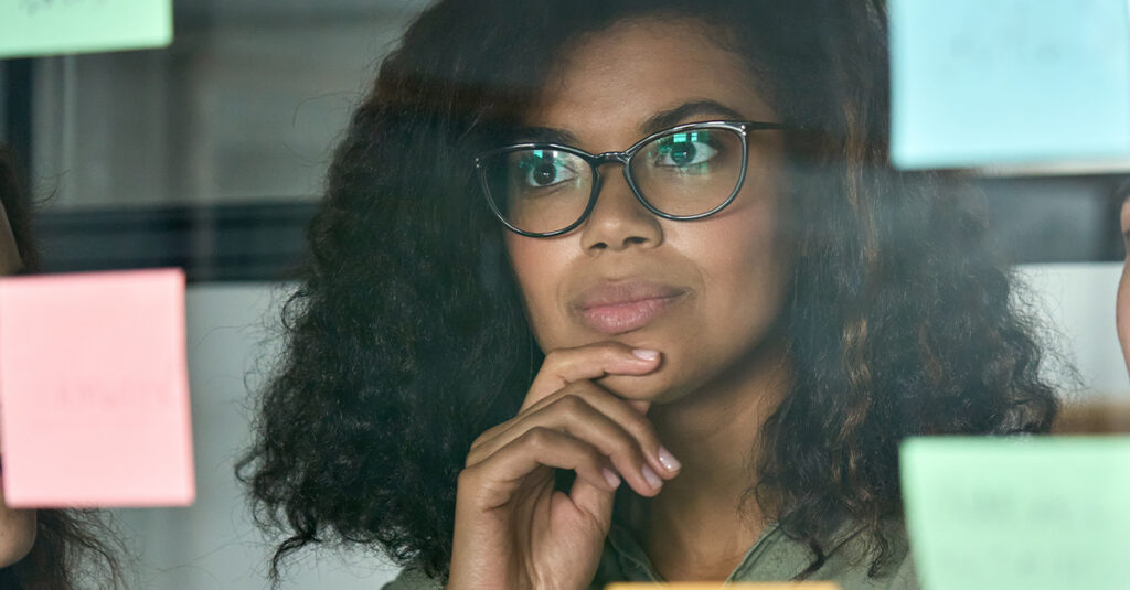 Young thoughtful African American woman in a modern office looking at adhesive sticky notes on a glass project board, shot through glass with reflection, illustrating the blog topic "12 Phrasal Verbs for the Corporate World"