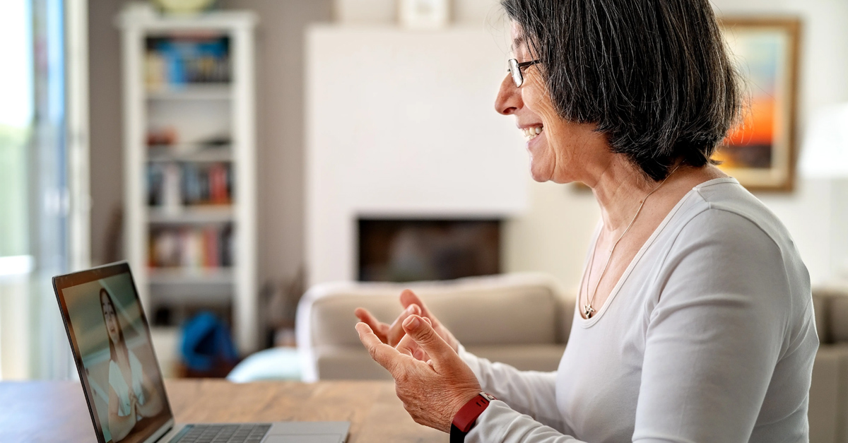 Side view of older woman smiling and interacting with a language class on her laptop in focus with her living room out of focus in the background.