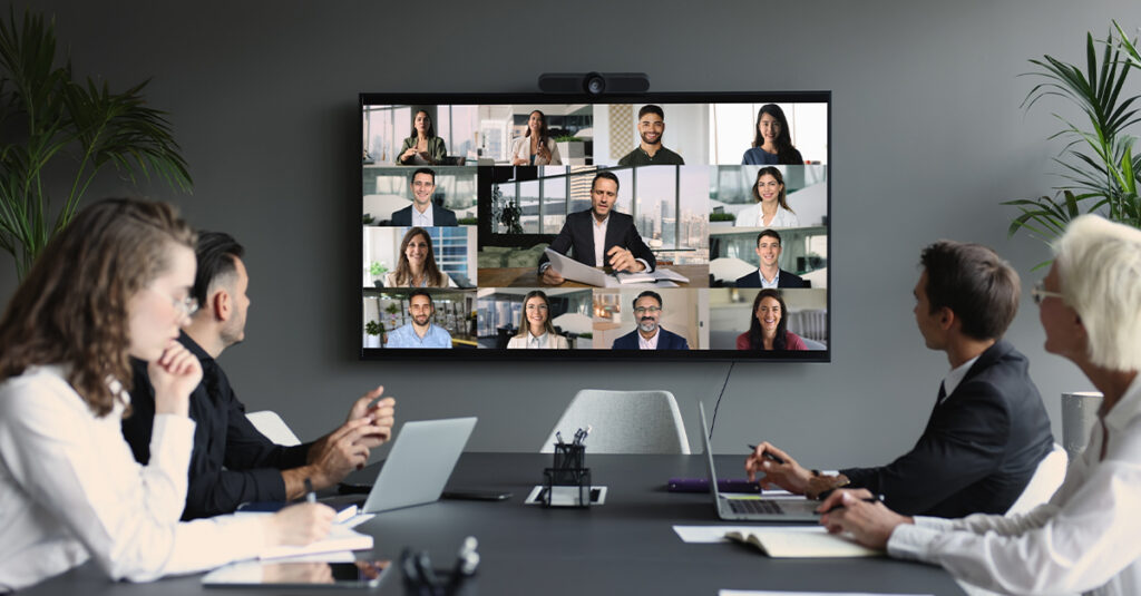 Group of diverse businesspeople gathered in boardroom engaging in video conference with multiple participants displayed on large screen.