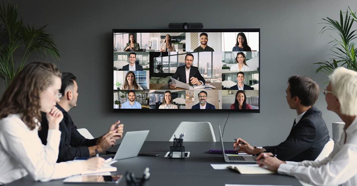 Group of diverse businesspeople gathered in boardroom engaging in video conference with multiple participants displayed on large screen.
