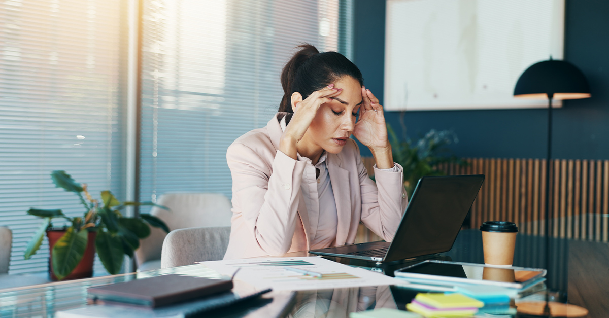 Professional woman at her desk in an office, hands on her temples while she stares at her laptop screen looking stressed