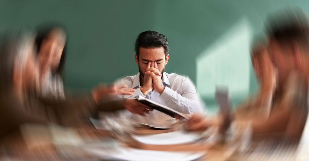 Man experiencing anxiety in a meeting in an office, with a blurred fish-eye effect on his coworkers while he closes his eyes and presses his hands to his face