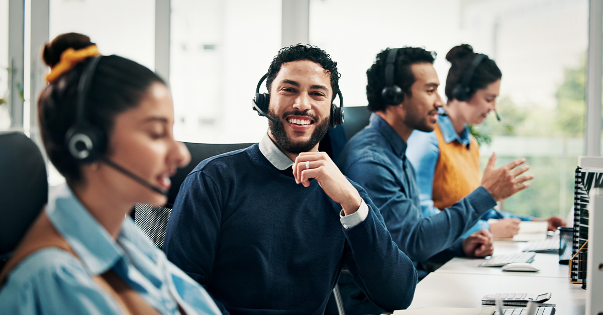 Customer support male employee in focus smiling and looking at the camera while three coworkers face their computers